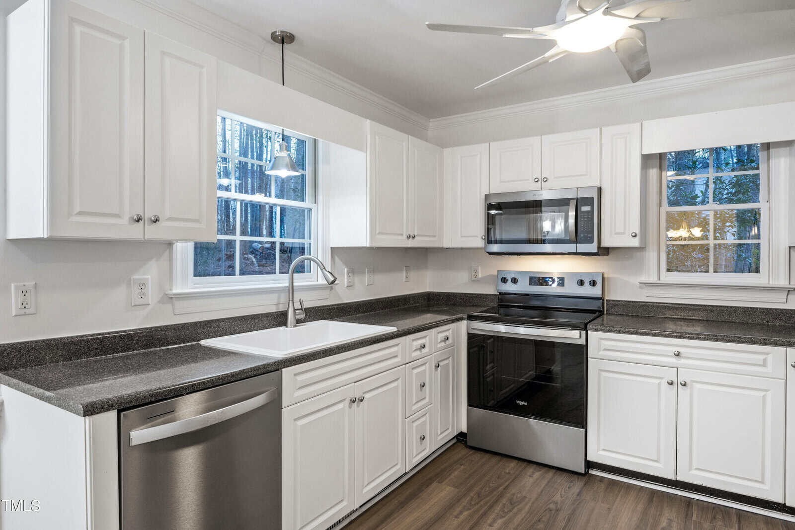 Modern kitchen with white cabinets, stainless steel appliances, dark countertops, a sink under a window, and wood-style flooring. The room is well-lit with natural light and a ceiling fan overhead.
