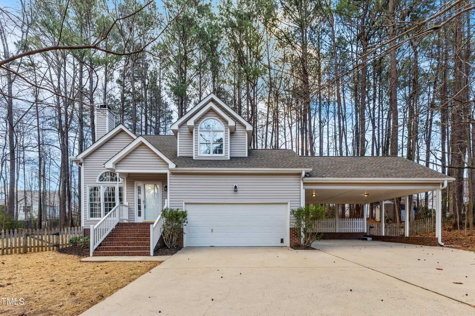 A two-story suburban house with gray siding, white trim, a front porch with brick steps, an attached two-car garage, a carport, and surrounded by trees on a concrete driveway.