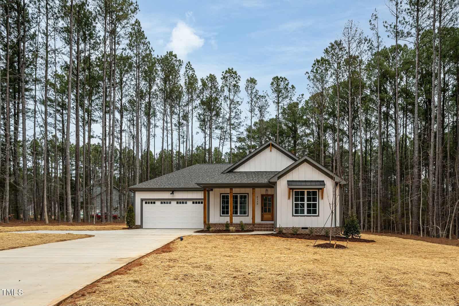 A single-story house with a gabled roof is nestled among tall trees. It features a white facade with wooden accents, a double garage, and a large driveway. The front yard is covered with straw. The sky is partly cloudy.