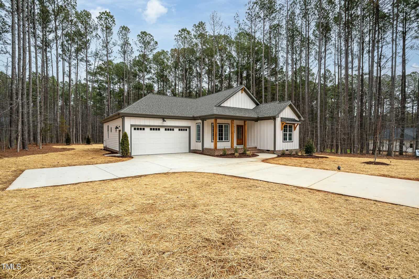 A single-story house with a gray roof and white siding is surrounded by tall pine trees. It features a two-car garage and a covered front porch. The driveway and walkway are newly paved, and the yard is covered with straw.