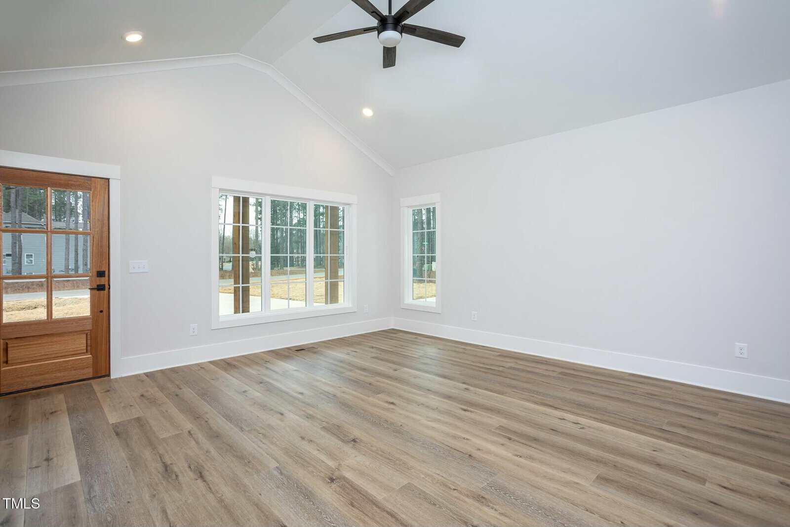 A spacious, unfurnished living room with light wood flooring and white walls. It features a high ceiling with a black ceiling fan, large windows offering a view outside, and a wooden entry door with glass panels.