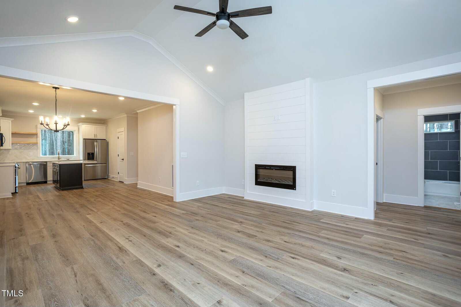 A spacious, empty living area with light wood flooring and white walls. There's a ceiling fan, a built-in electric fireplace on the right wall, and an open doorway leading to a modern kitchen with stainless steel appliances and a chandelier.