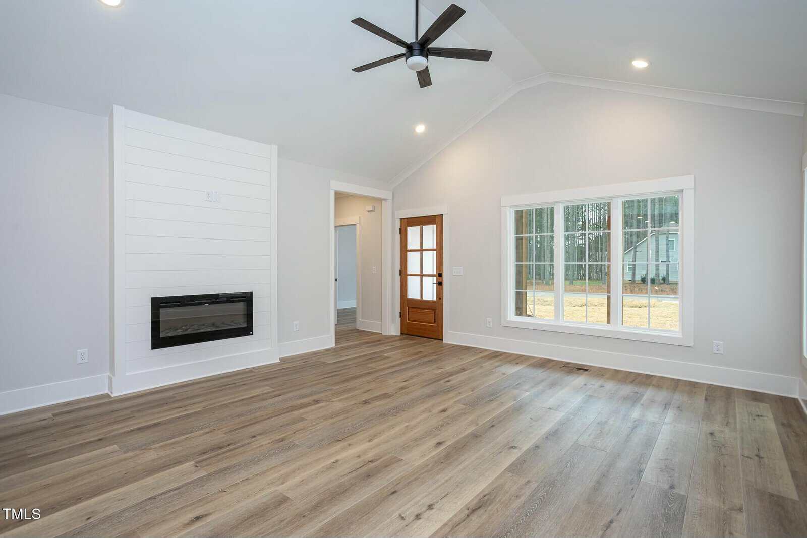 A spacious living room with light wood flooring, white walls, and a vaulted ceiling. It features a modern electric fireplace on the left and a large window on the right. A wooden door with glass panels is in the center. Ceiling lights and a fan are present.
