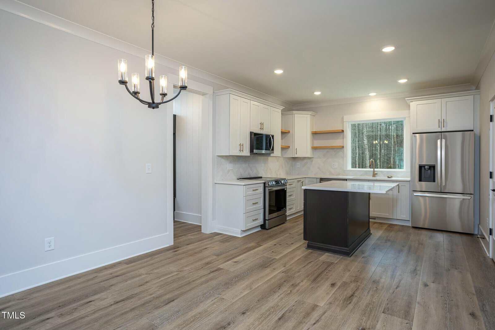 Modern kitchen with light wood flooring and white cabinets. Features include a black kitchen island, stainless steel appliances, a window above the sink, and a black chandelier. Walls are painted in a light neutral color.