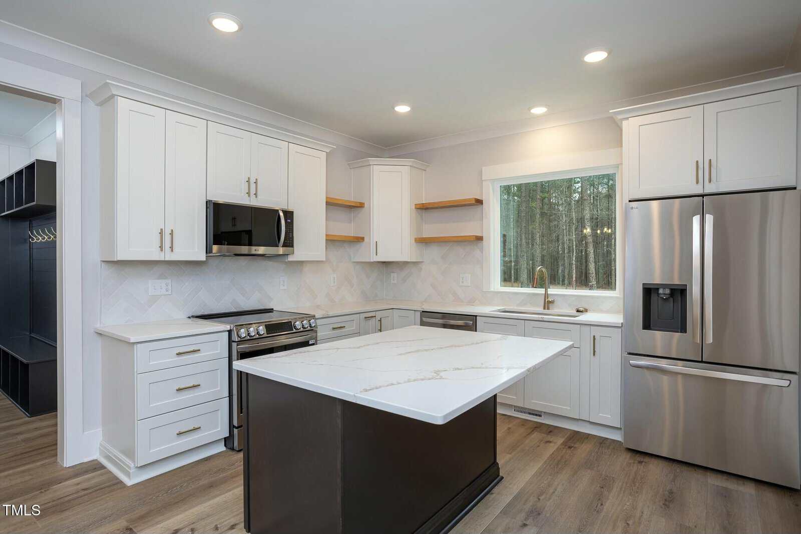 A modern kitchen with white cabinets, stainless steel appliances, and a central island with a marble countertop. The space features a large window, recessed lighting, and wooden shelves.