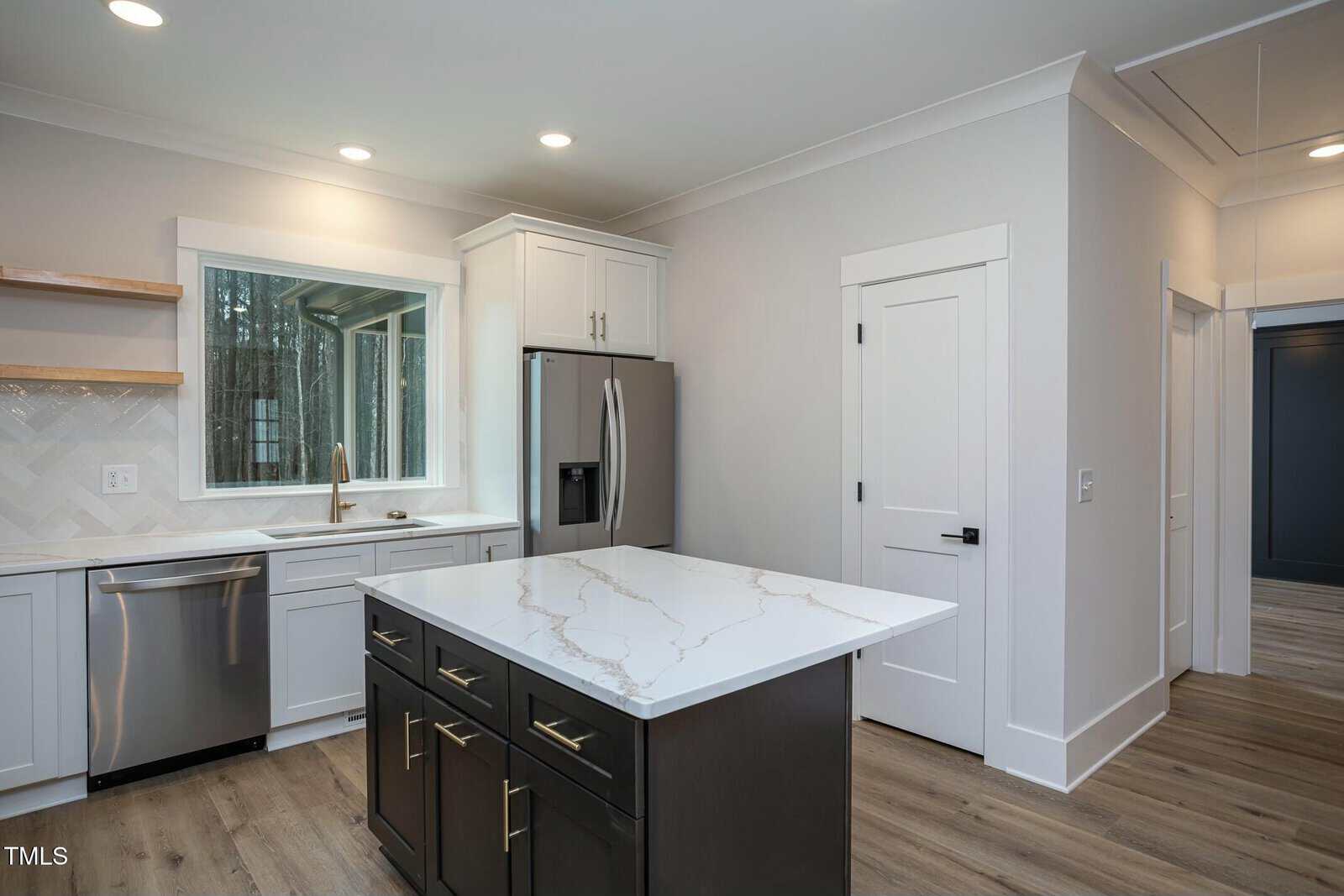 A modern kitchen featuring a marble-topped island with dark cabinetry, stainless steel refrigerator, and dishwasher. A large window above the sink provides natural light. The flooring is a light wood, and the walls are painted a light gray.
