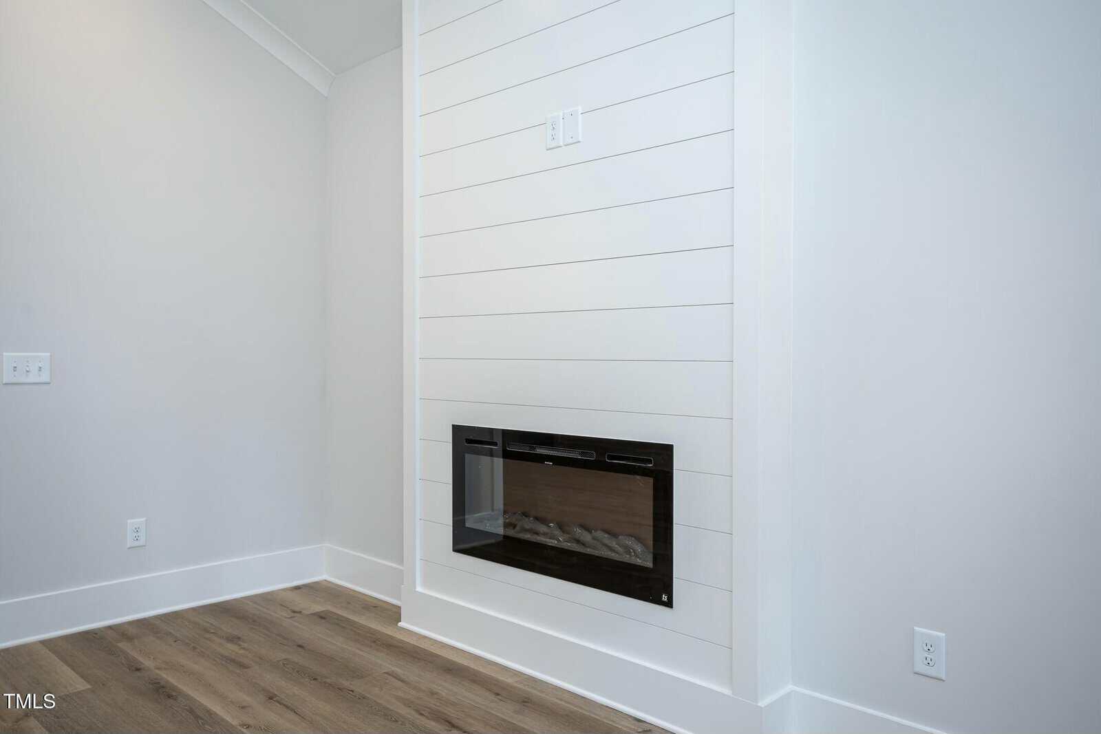 Modern room corner featuring a white shiplap wall with an electric fireplace insert. The floor is light wood, and the walls are painted white. An electrical outlet and light switch are visible on the left side.