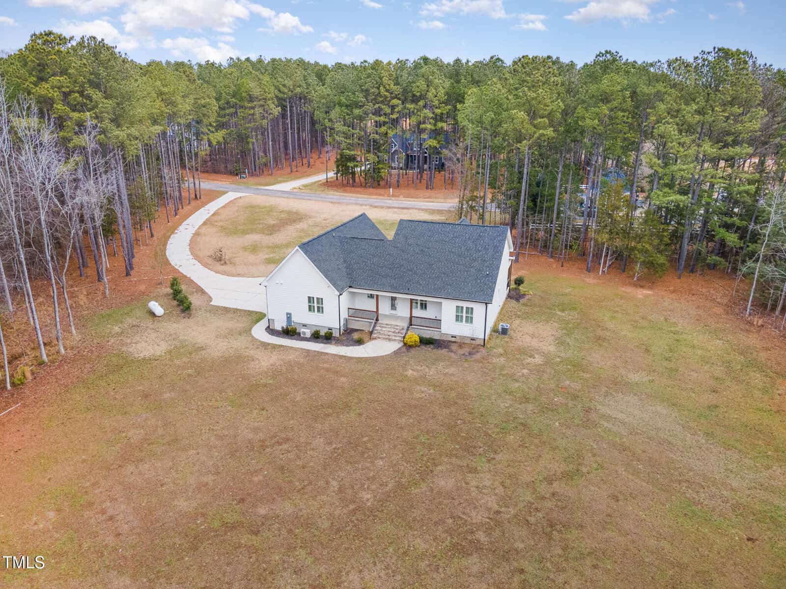 Aerial view of a single-story white house with a dark roof, surrounded by an expansive lawn and a pine forest. A curved driveway leads to the front of the house, with a few small shrubs lining the entrance. A road is visible in the background.
