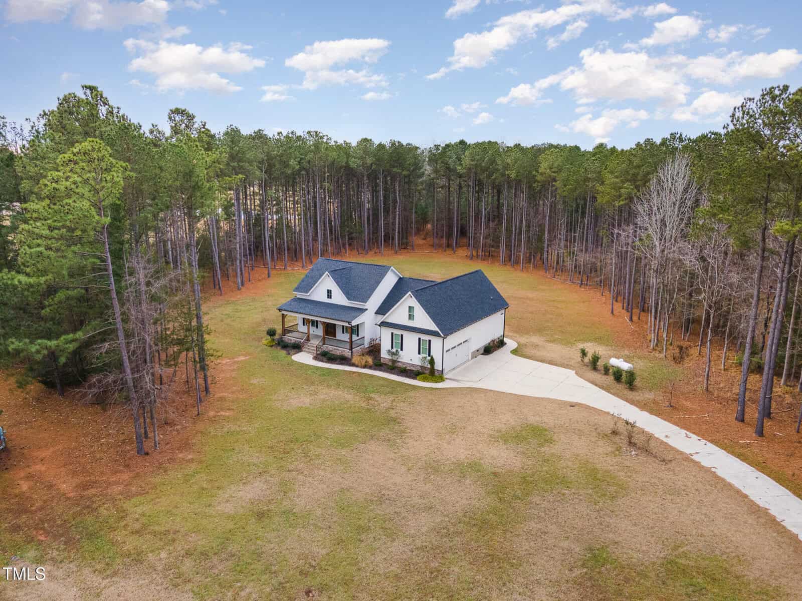 A white house with a dark roof sits on an expansive lawn surrounded by tall pine trees. The driveway curves to the front of the house, and the sky is partly cloudy. The scene appears to be in a rural setting.