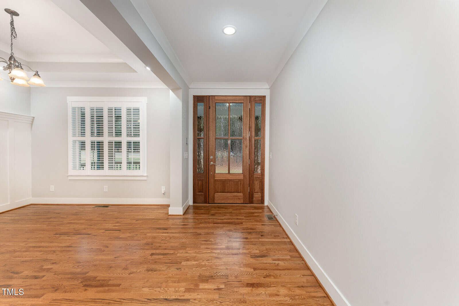 A room with polished hardwood floors, a wooden double door with glass panels, and a white interior featuring a window with shutters. The ceiling has recessed lighting, and the overall decor is minimalist.