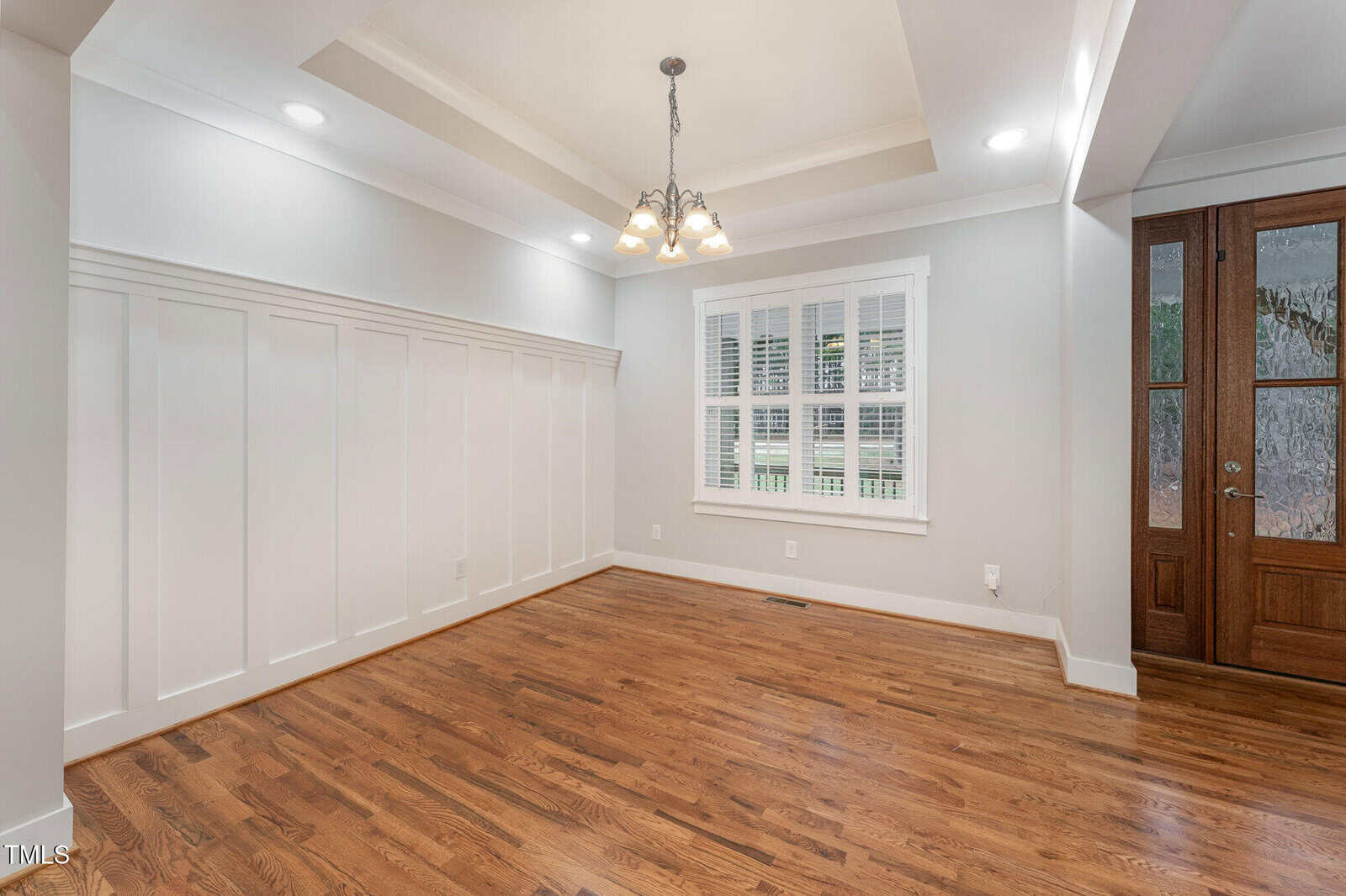 An empty room with hardwood floors and white wainscoting on the walls. A window with white blinds is on one wall. A wooden door with glass panels is to the right. A chandelier with multiple bulbs hangs from the ceiling.