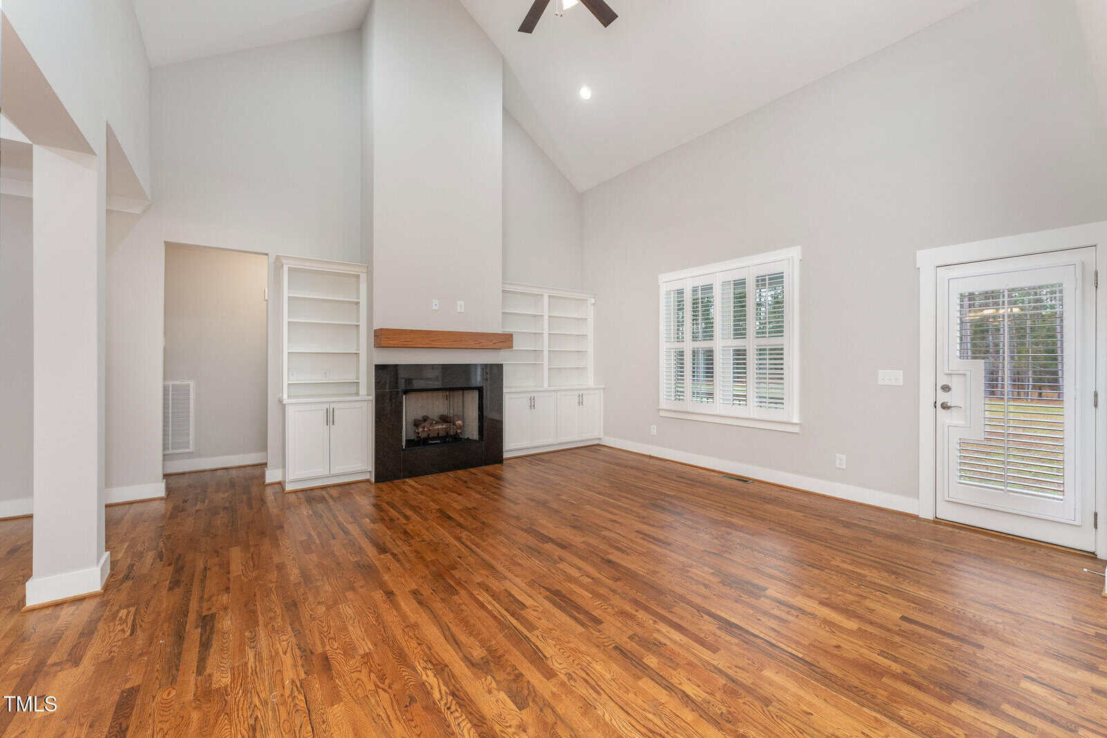 Spacious living room with high vaulted ceilings, hardwood floors, and a modern fireplace. Large windows and a glass door provide natural light. Built-in shelving units flank the fireplace, and the walls are painted in light neutral colors.