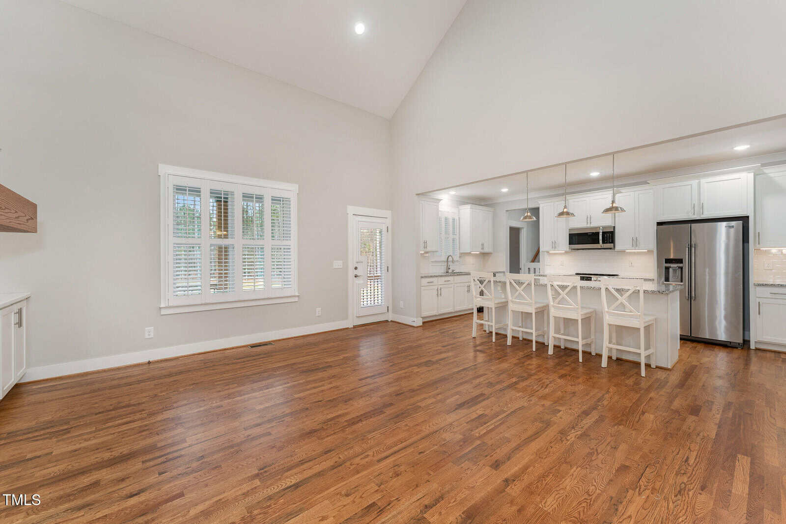 An open-concept kitchen and living area with hardwood floors. The kitchen features white cabinets, a stainless steel refrigerator, and a breakfast bar with barstools. The room is lit by recessed lighting and pendant lights, with a window and a door on the left.