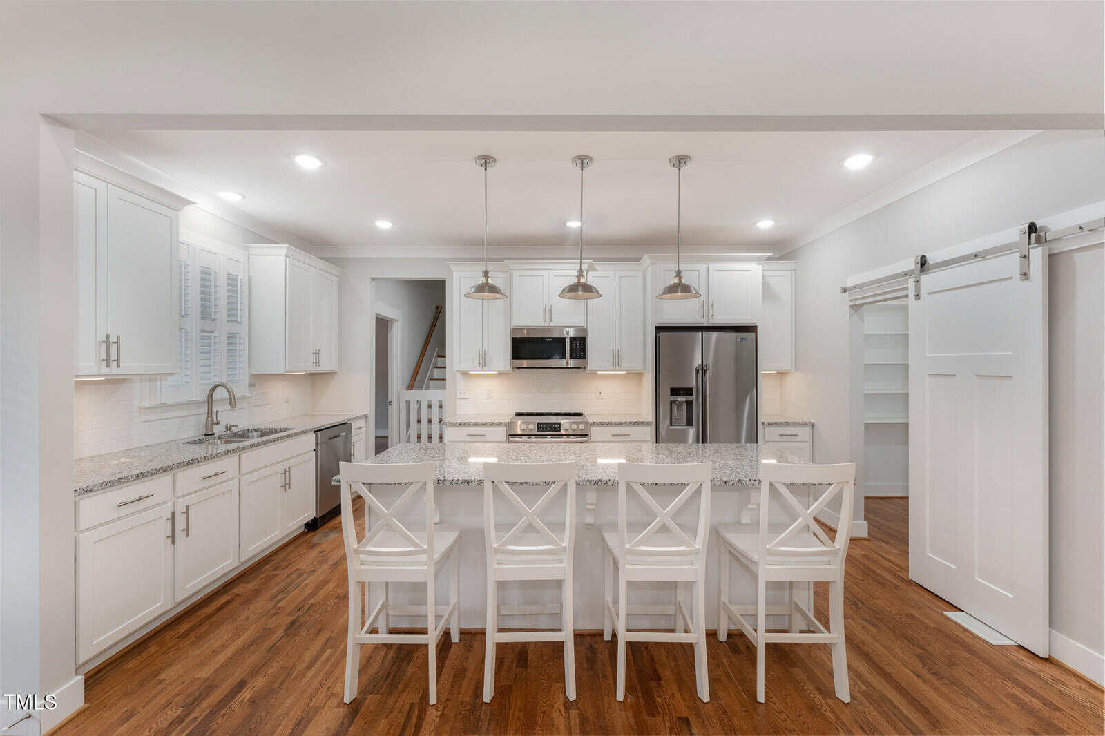 A modern kitchen with white cabinetry and a central island featuring a granite countertop. Four white bar stools are aligned at the island. Stainless steel appliances include a refrigerator and oven. Pendant lights hang above the island, and wooden floors are visible.