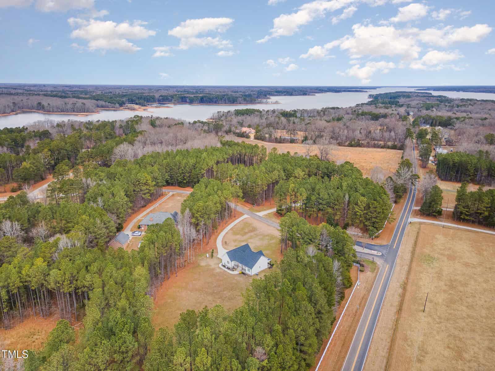 Aerial view of a rural landscape with a winding road, surrounded by dense green forest and a few houses. In the background, a large body of water is visible under a partly cloudy sky.