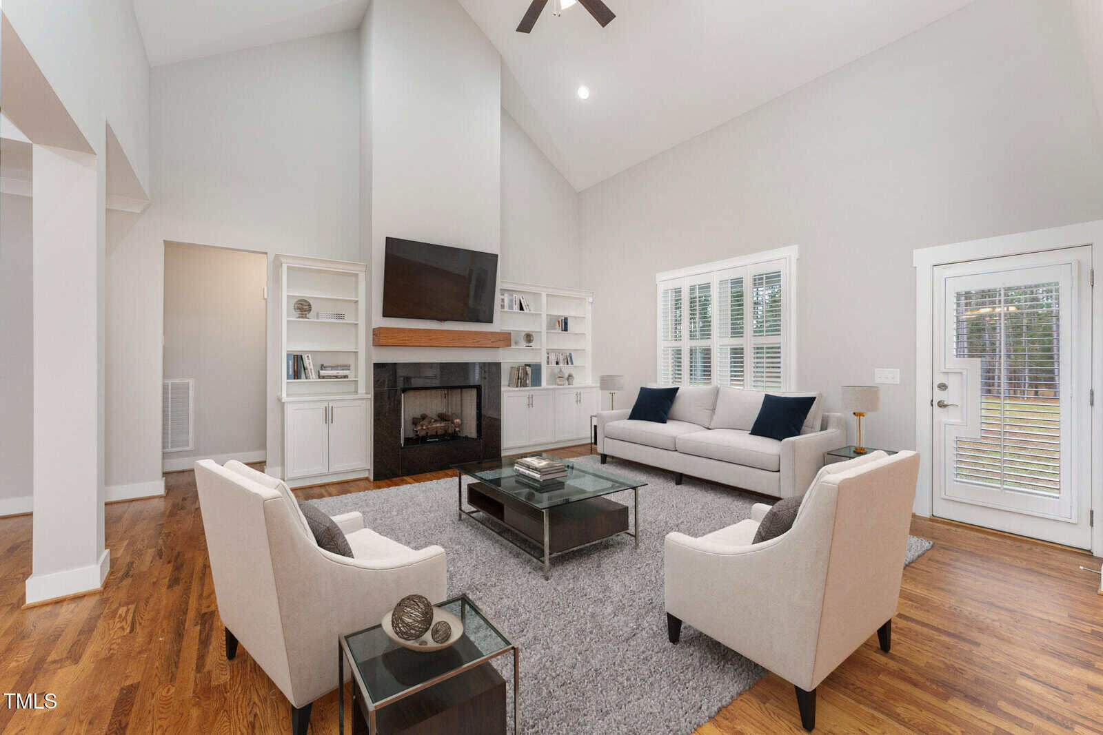 A modern living room with light-toned walls features a fireplace, a mounted TV, and built-in shelves. A gray sofa and two armchairs are arranged around a glass coffee table on a gray rug. Large windows and a glass door let in natural light.