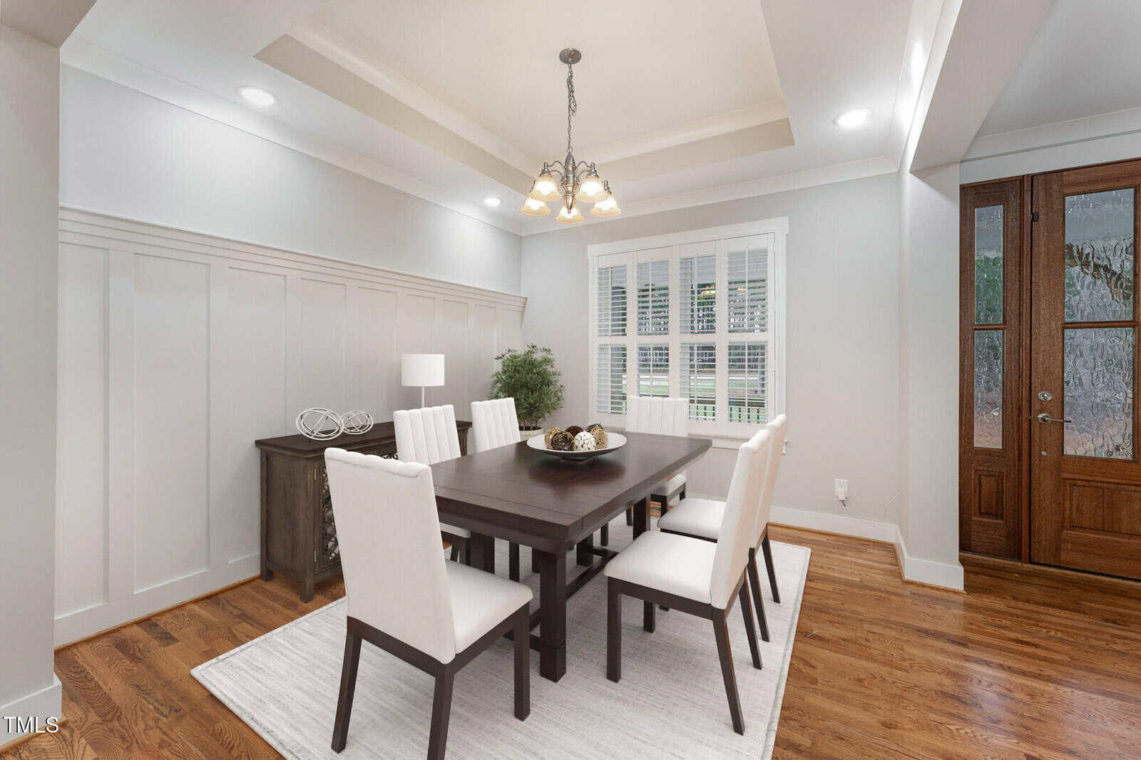 Dining room featuring a dark wooden table with six upholstered chairs on a light rug. A modern chandelier hangs above. A sideboard with decor items is against the wall, and a window with blinds is visible. The entrance shows a wooden door with glass panels.