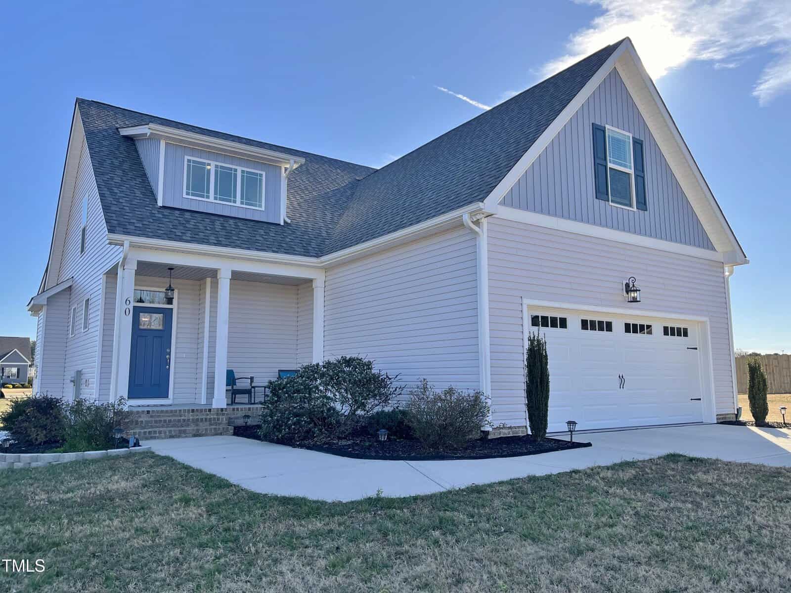 A two-story suburban house with light gray siding, a front porch, blue front door, double garage, small shrubs, and a manicured lawn under a clear blue sky.