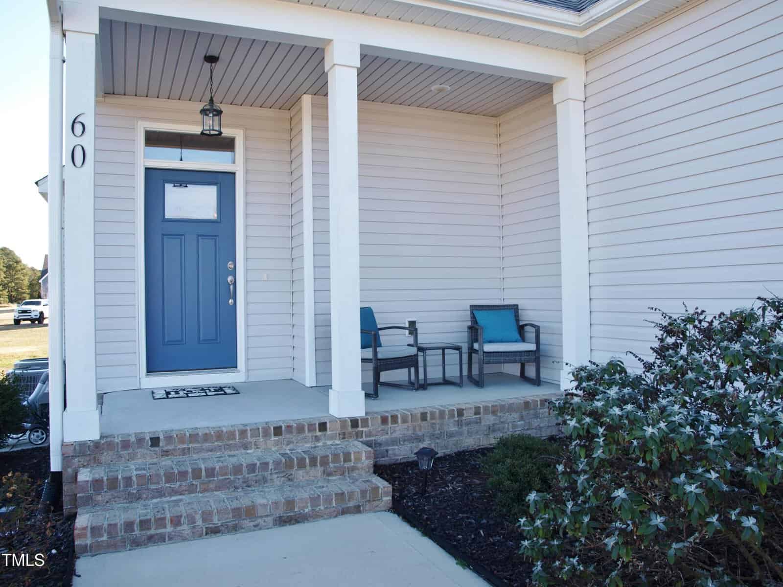 A front porch with brick steps, a blue door, white siding, and two chairs with cushions next to a small table. A hanging light fixture is above the door, and bushes line the walkway. House number 60 is visible.