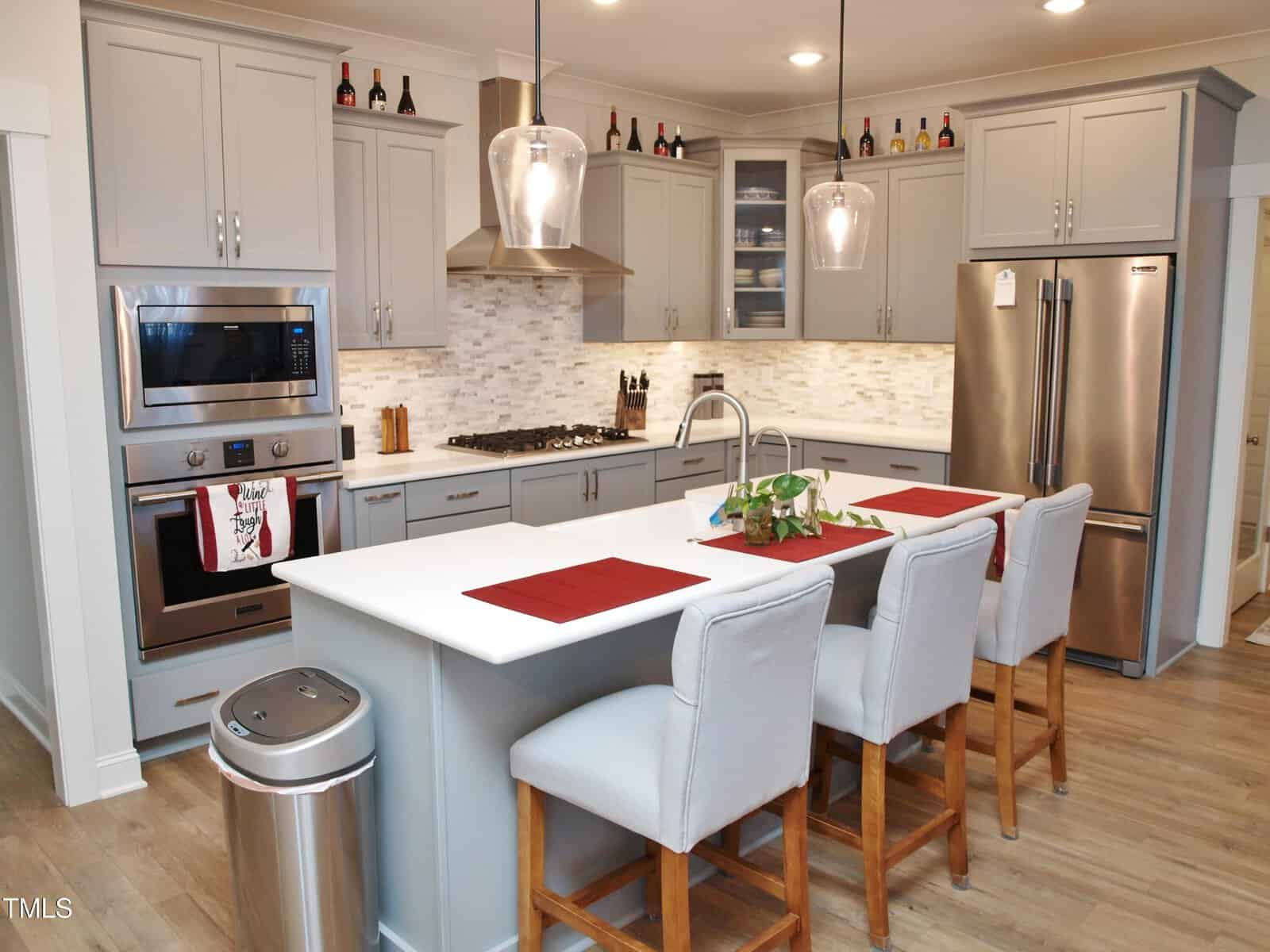 A modern kitchen with gray cabinets, stainless steel appliances, a center island with a sink, three upholstered chairs, pendant lights, and wine bottles displayed above the cabinets. Red placemats are on the island.