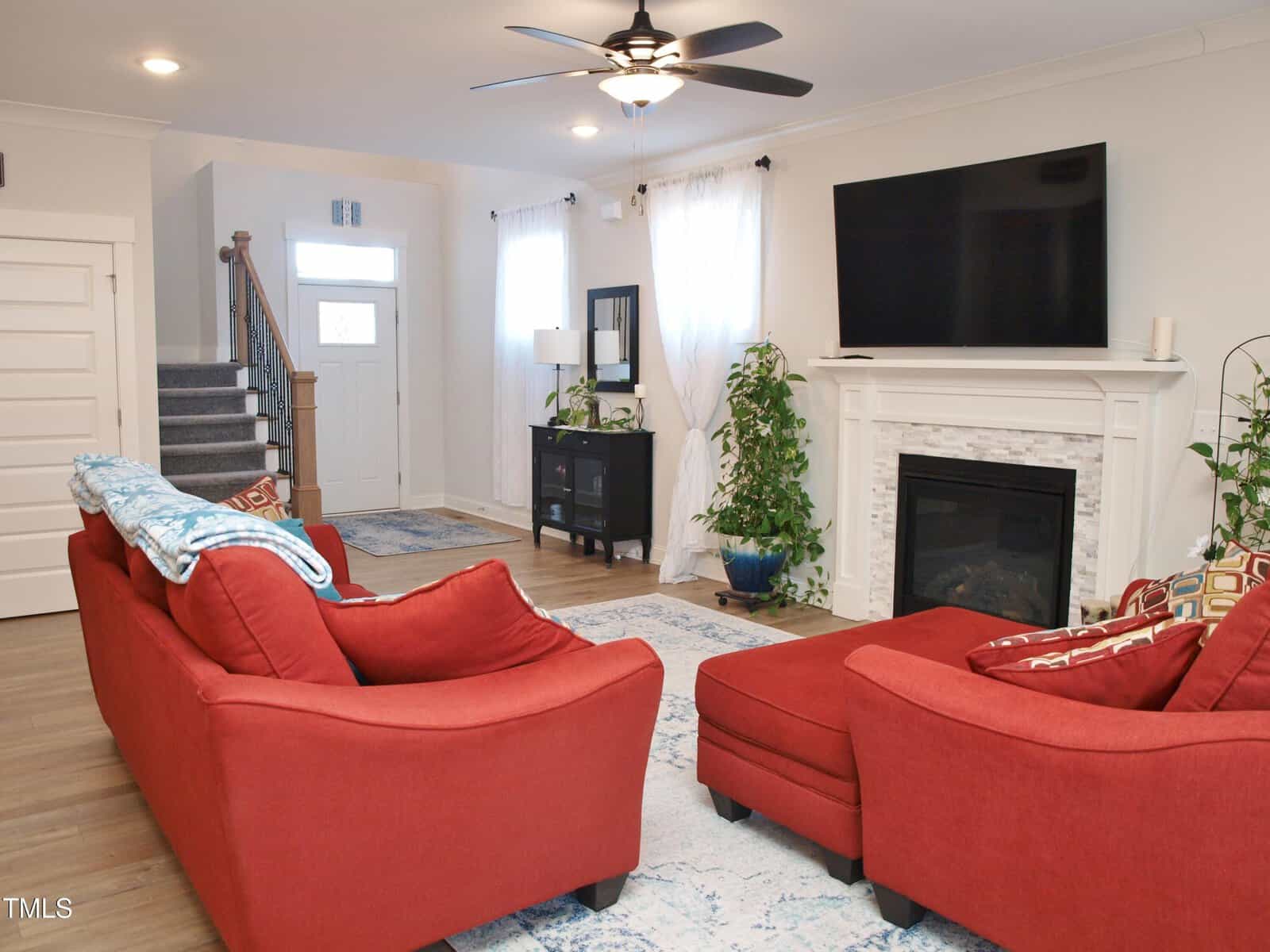 A bright living room with red sofas and matching ottoman, a white fireplace, wall-mounted TV, potted plants, and light blue accents. Stairs and a front door are visible in the background.