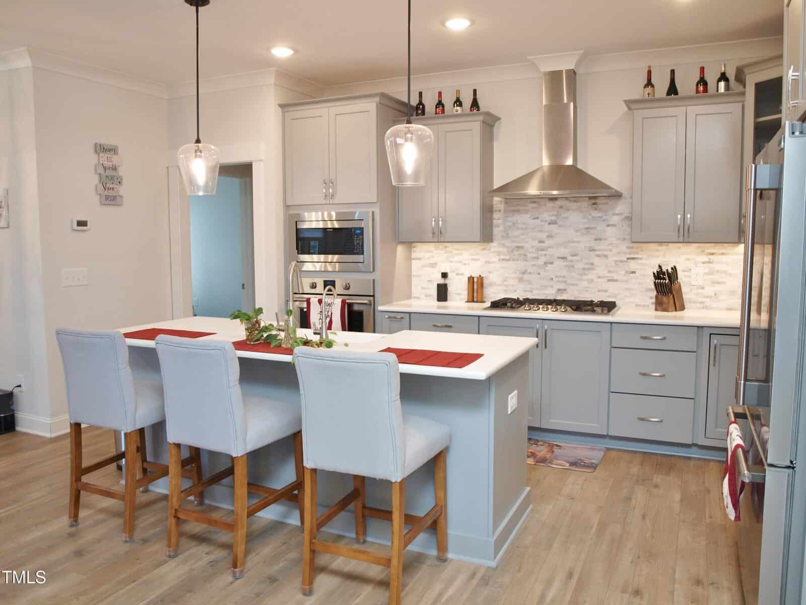 Modern kitchen with gray cabinets, stainless steel appliances, a white island with three upholstered barstools, pendant lights, and a tile backsplash. Decorative wine bottles and art hang above the cabinets.