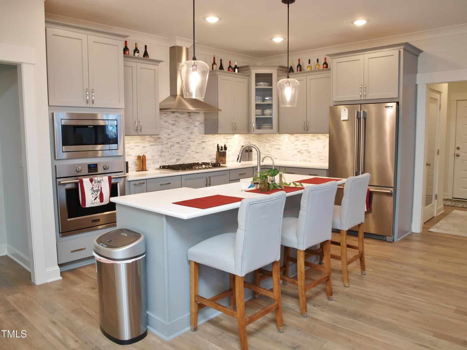 Modern kitchen with gray cabinets, stainless steel appliances, pendant lights, and an island with three upholstered chairs. Wine bottles are displayed above the cabinets, and the counters have minimal decor.