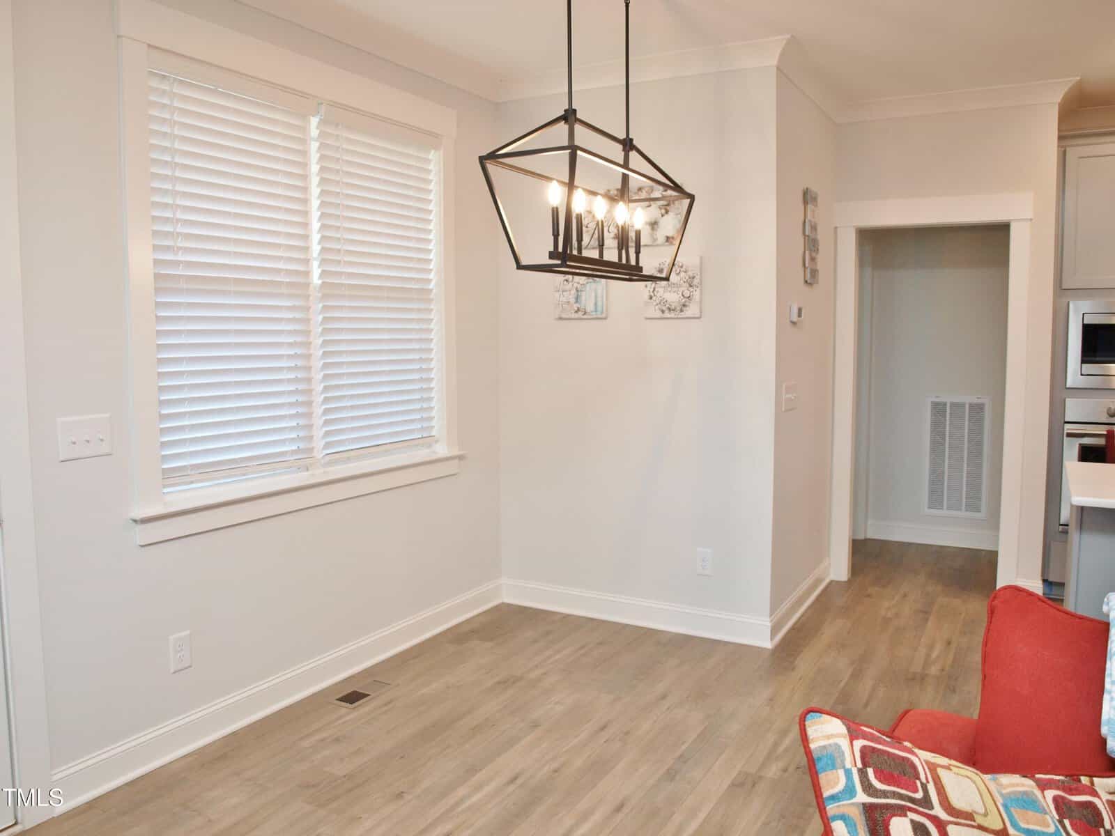 Modern dining area with light wood flooring, a geometric black chandelier, white blinds on the window, neutral walls, and a partial view of a red chair with colorful pillows in the lower right corner.