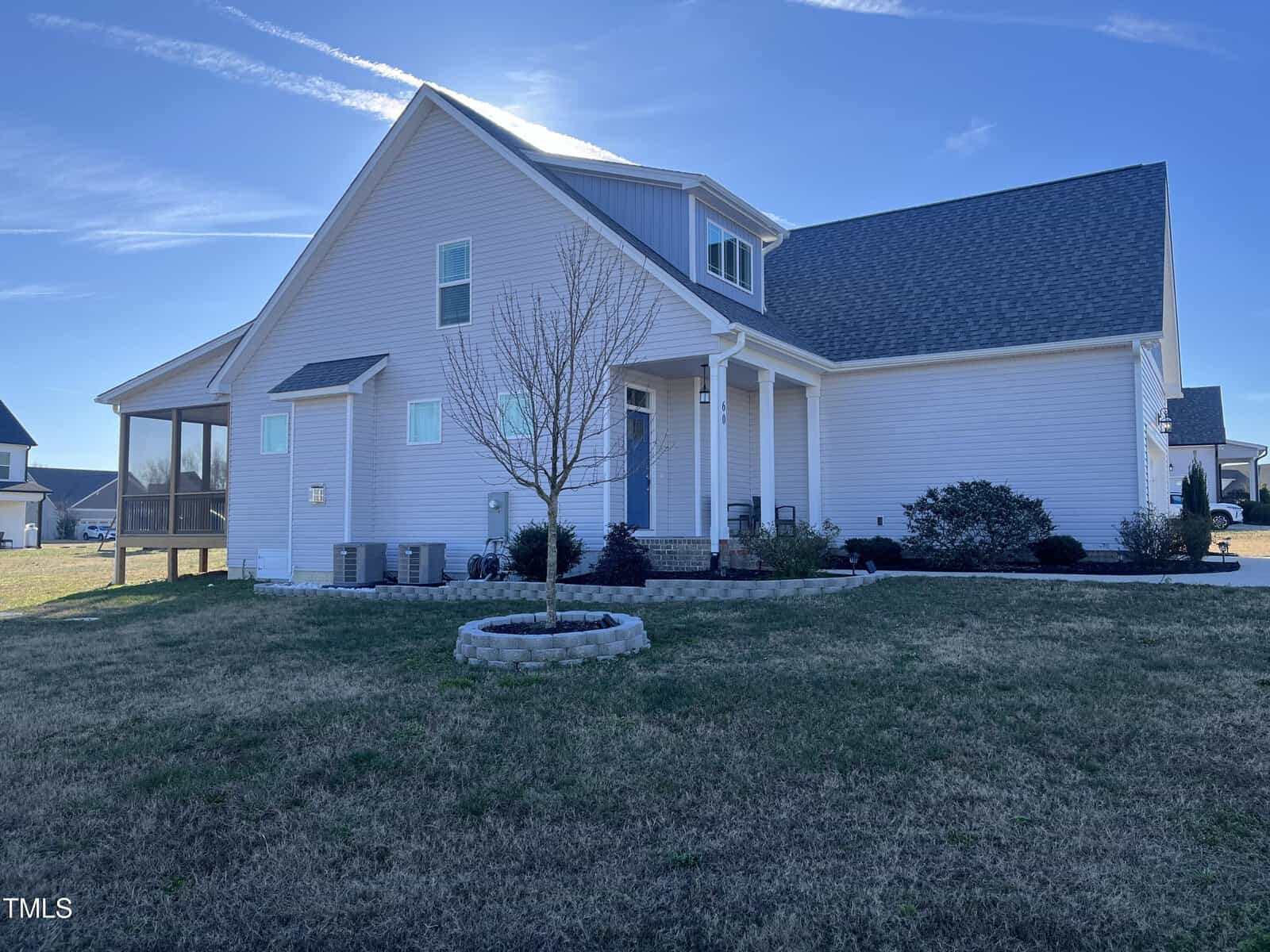 Single-story light-colored house with a gabled roof, front porch, and screened-in side patio. A small leafless tree is planted in a circular stone bed in the front yard, with trimmed bushes near the house.