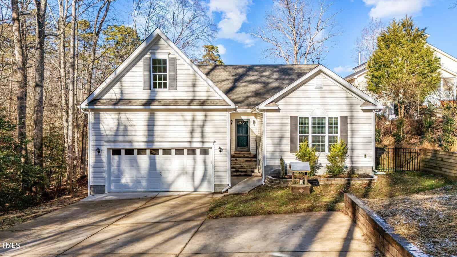 A light-colored, single-story house with a two-car garage, front steps leading to the entrance, and a small landscaped yard, surrounded by trees and a wooden fence, under a blue sky.