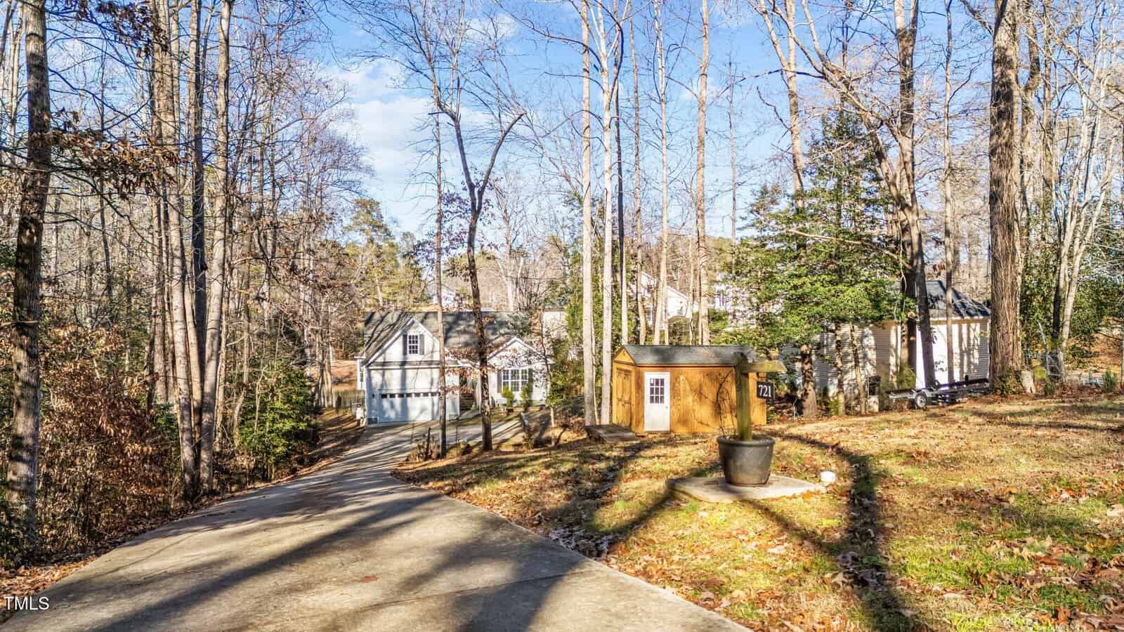 A concrete driveway leads to a house with a two-car garage, surrounded by tall, leafless trees. A small shed and a large black flower pot sit on the right side of the driveway. The scene is well-lit with sunlight.