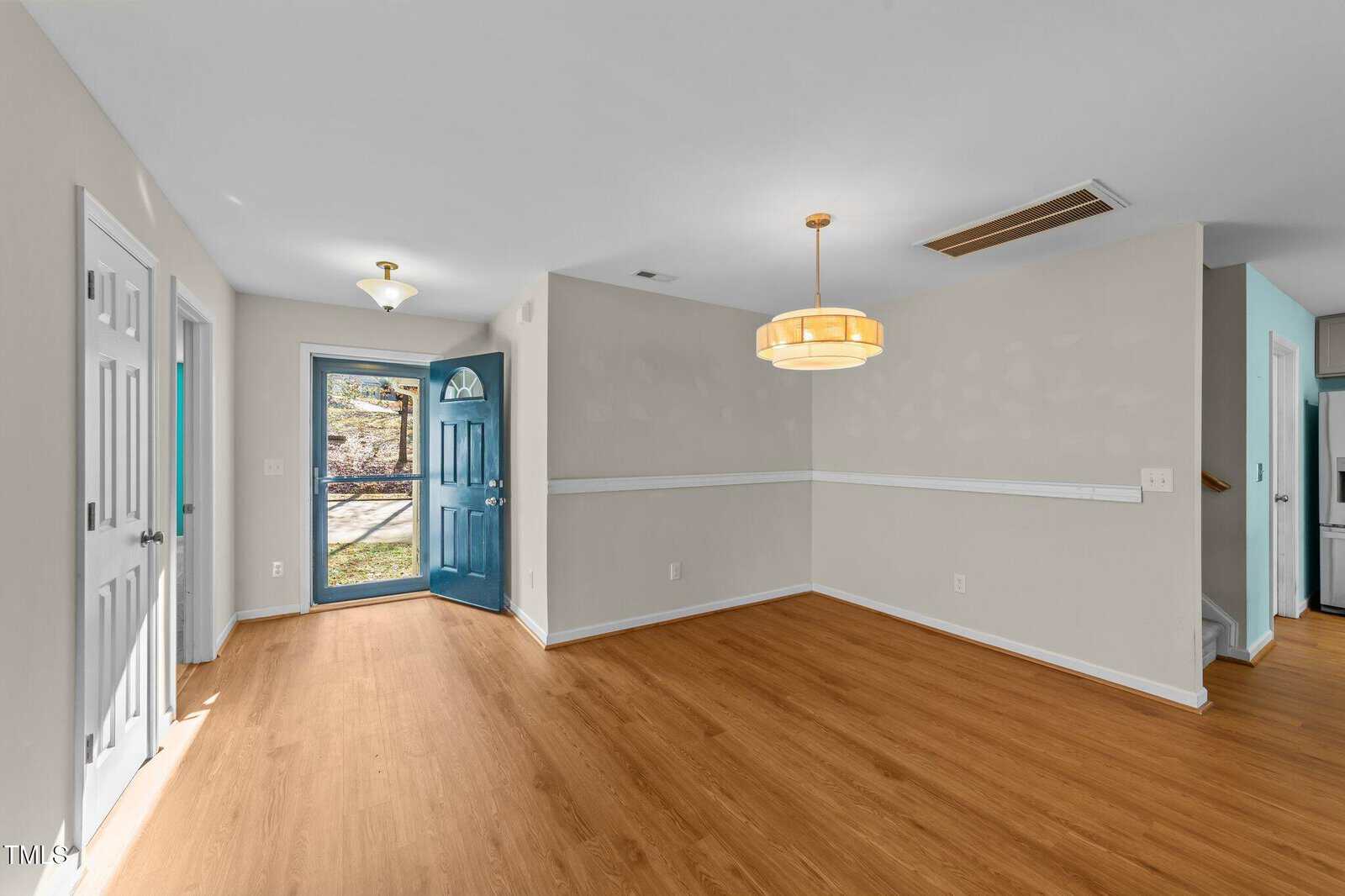 A room with light wood flooring, beige walls, a blue front door, and a round ceiling light fixture. There are two white doors on the left and an entrance to another room on the right. Natural light enters from the front door.
