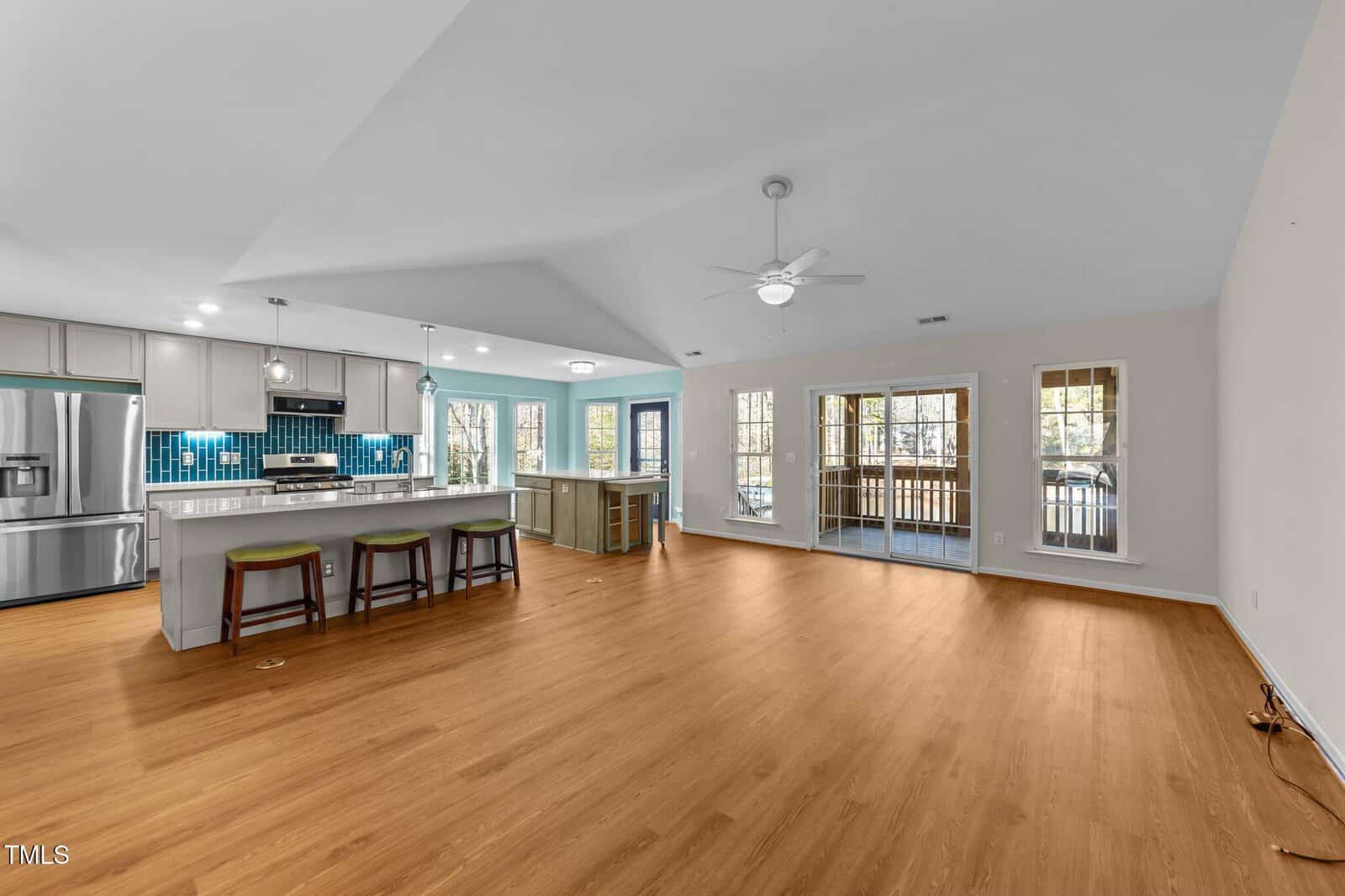 Spacious open-concept living area with wood flooring, featuring a modern kitchen with stainless steel appliances, blue tile backsplash, an island with barstools, and large sliding glass doors leading to a screened porch.