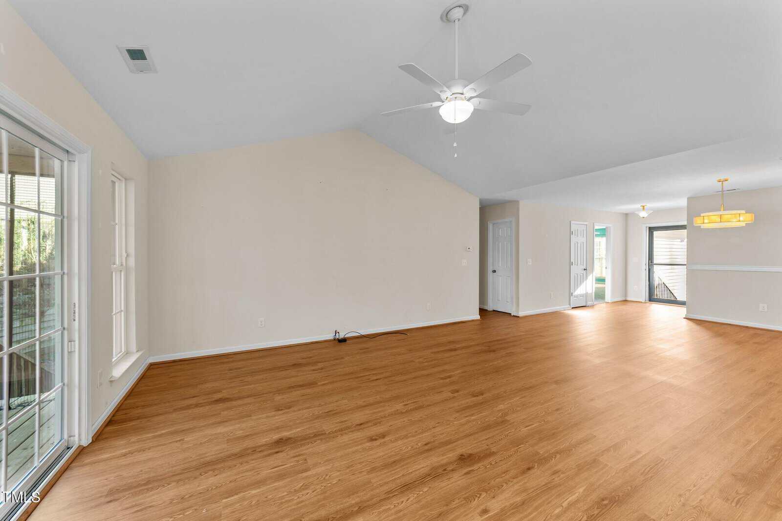 A spacious, empty living and dining area with light wooden floors, beige walls, vaulted ceiling with a ceiling fan, large windows, sliding glass door, and a modern light fixture above the dining space.