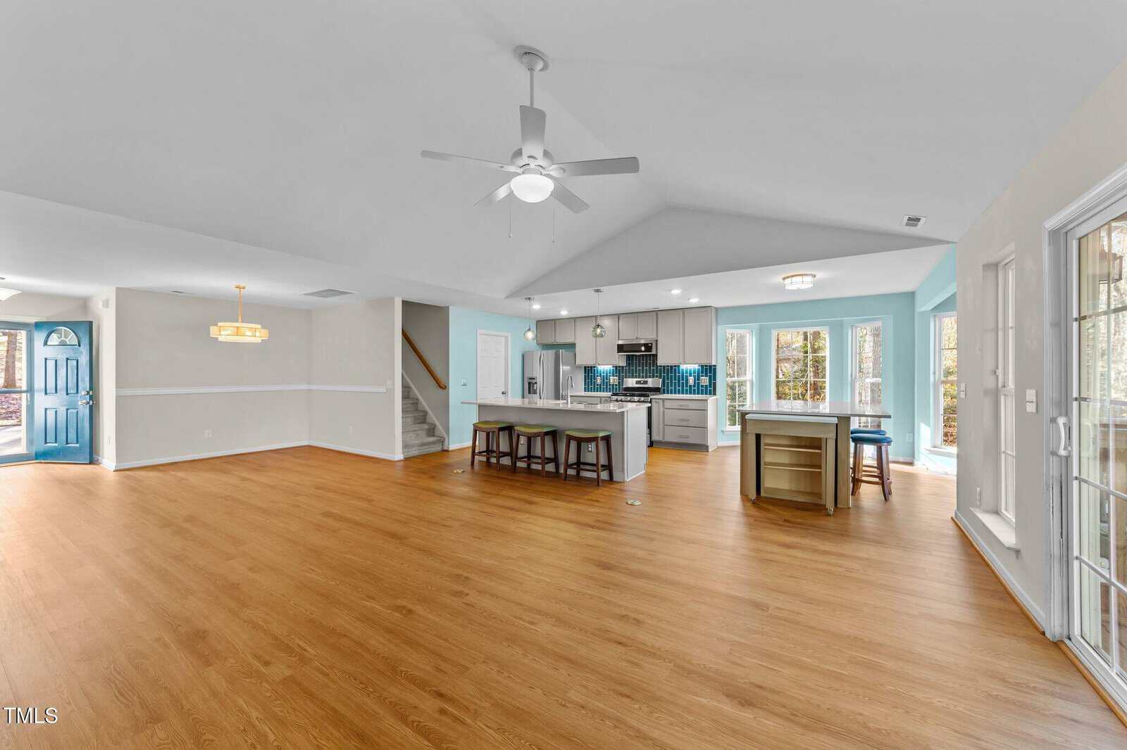 Open-concept kitchen and living area with light wood floors, blue and white walls, ceiling fan, two islands, stainless steel appliances, and large windows letting in natural light. Stairs and a blue front door are visible.