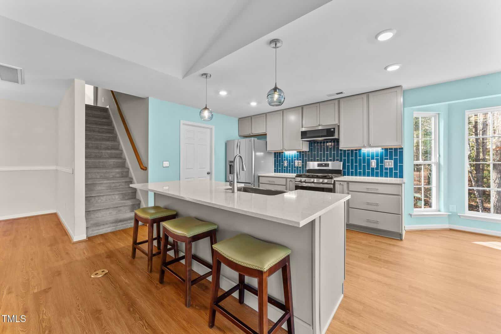 Modern kitchen with a large island, three green-cushioned stools, light gray cabinets, blue tile backsplash, stainless steel appliances, and wood floors, next to a staircase and large windows with natural light.