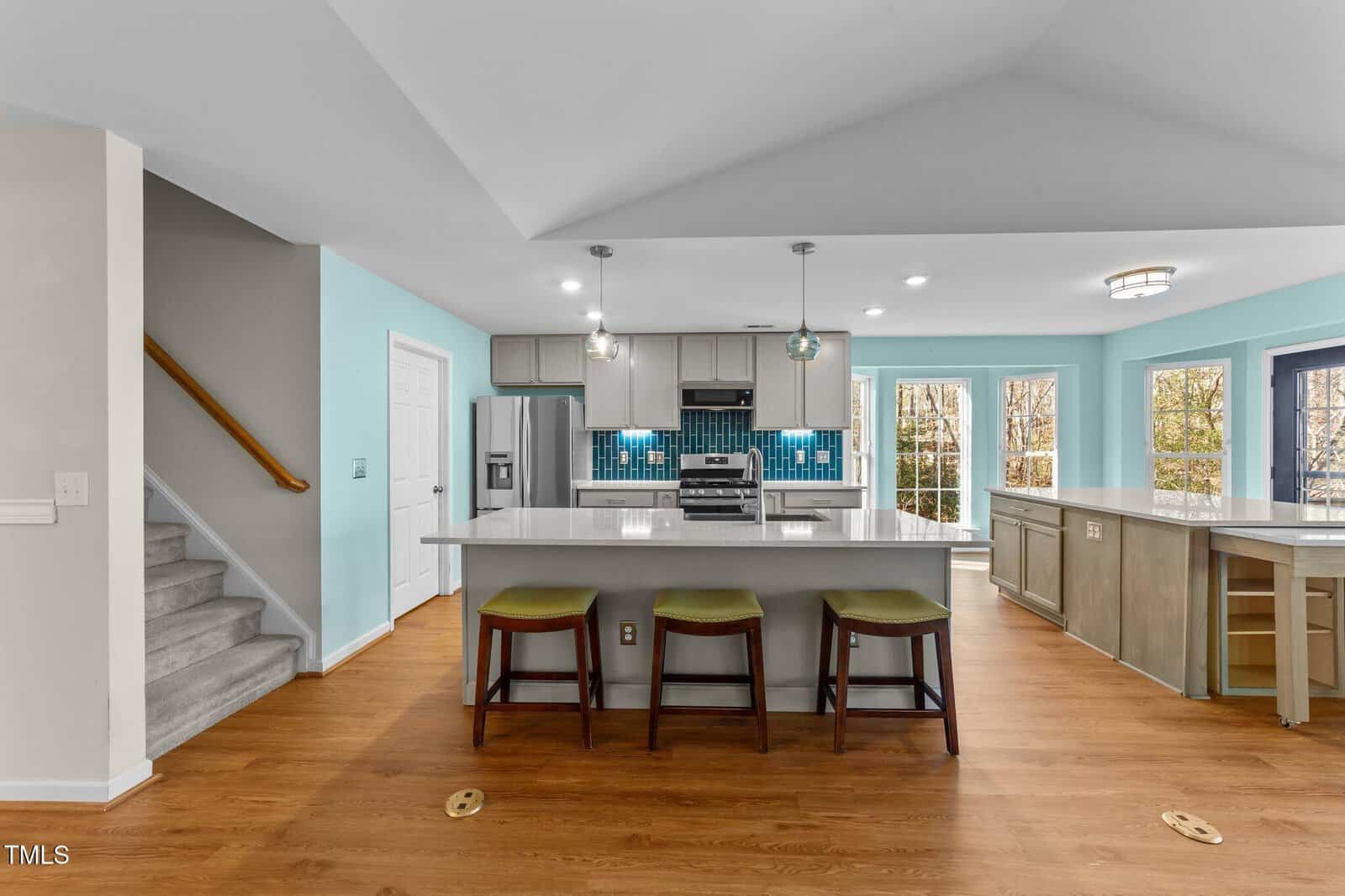 Modern kitchen with island, three stools, stainless appliances, blue tile backsplash, light wood floors, and light blue walls. Staircase on the left, large windows and glass door on the right.
