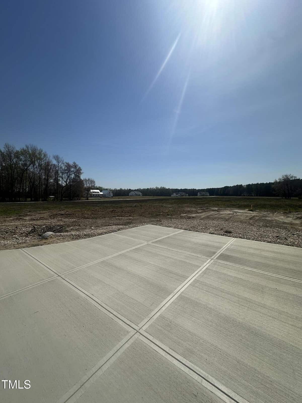 A concrete driveway extends into a flat, open plot of land with sparse grass and trees in the background under a clear, sunny sky. A few houses are visible in the distance.