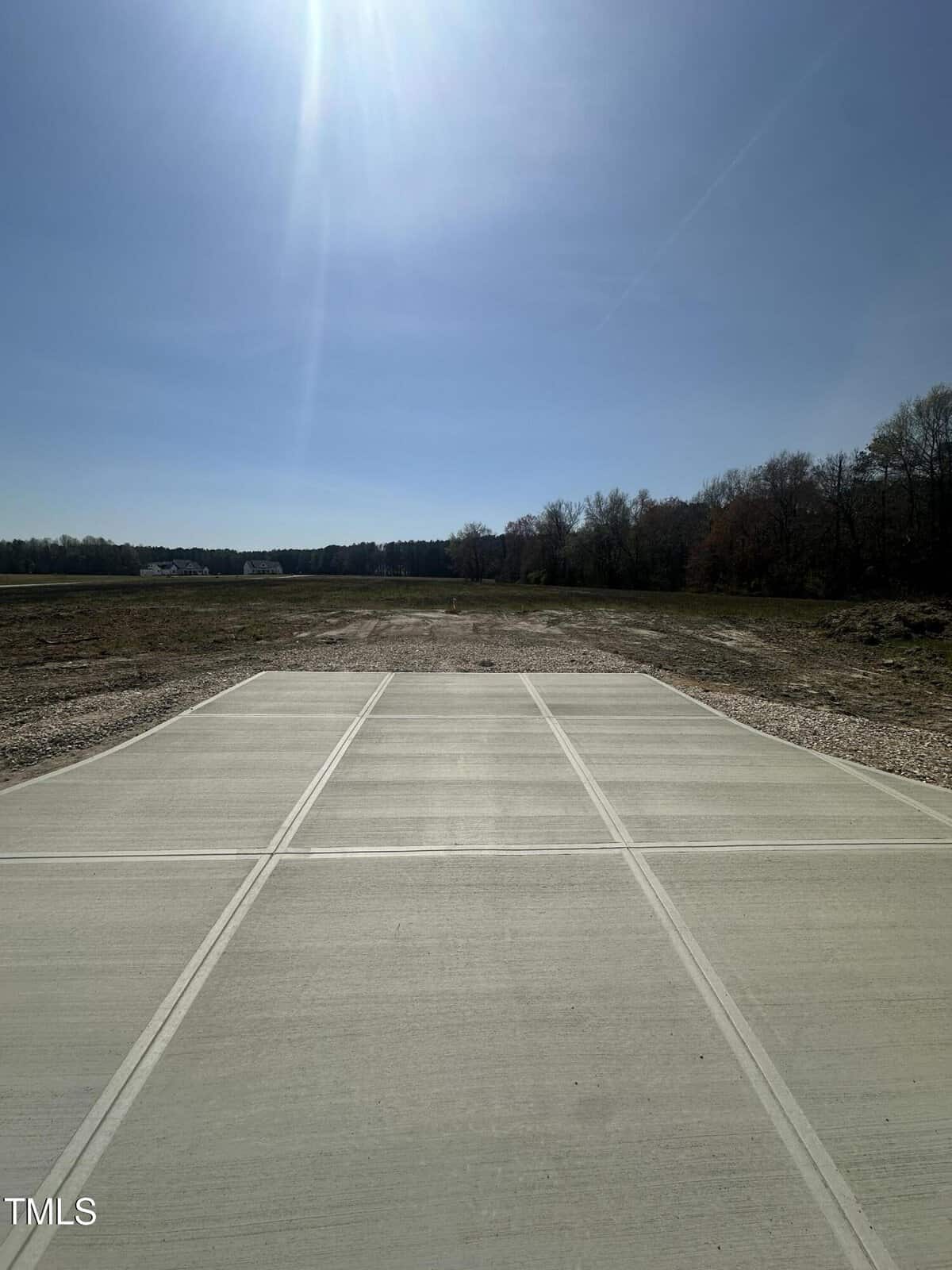 A rectangular concrete slab occupies the foreground, leading to a flat, open field with sparse grass. Trees border the area on the right, and the sky above is clear and sunny.