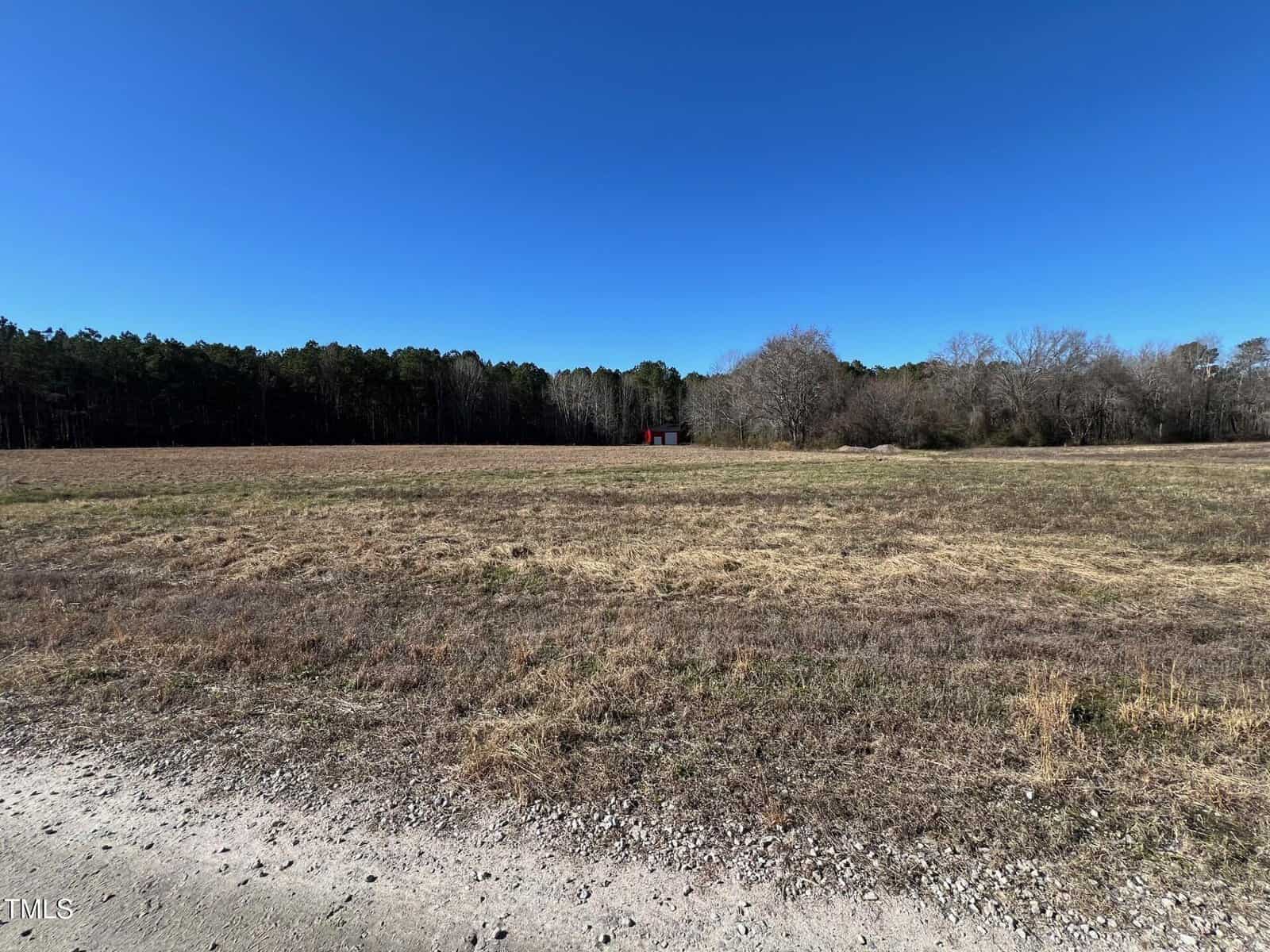 A wide, open field with dry grass under a clear blue sky. Trees line the horizon in the background, and a small red structure is visible near the center right. A gravel path runs along the foreground.