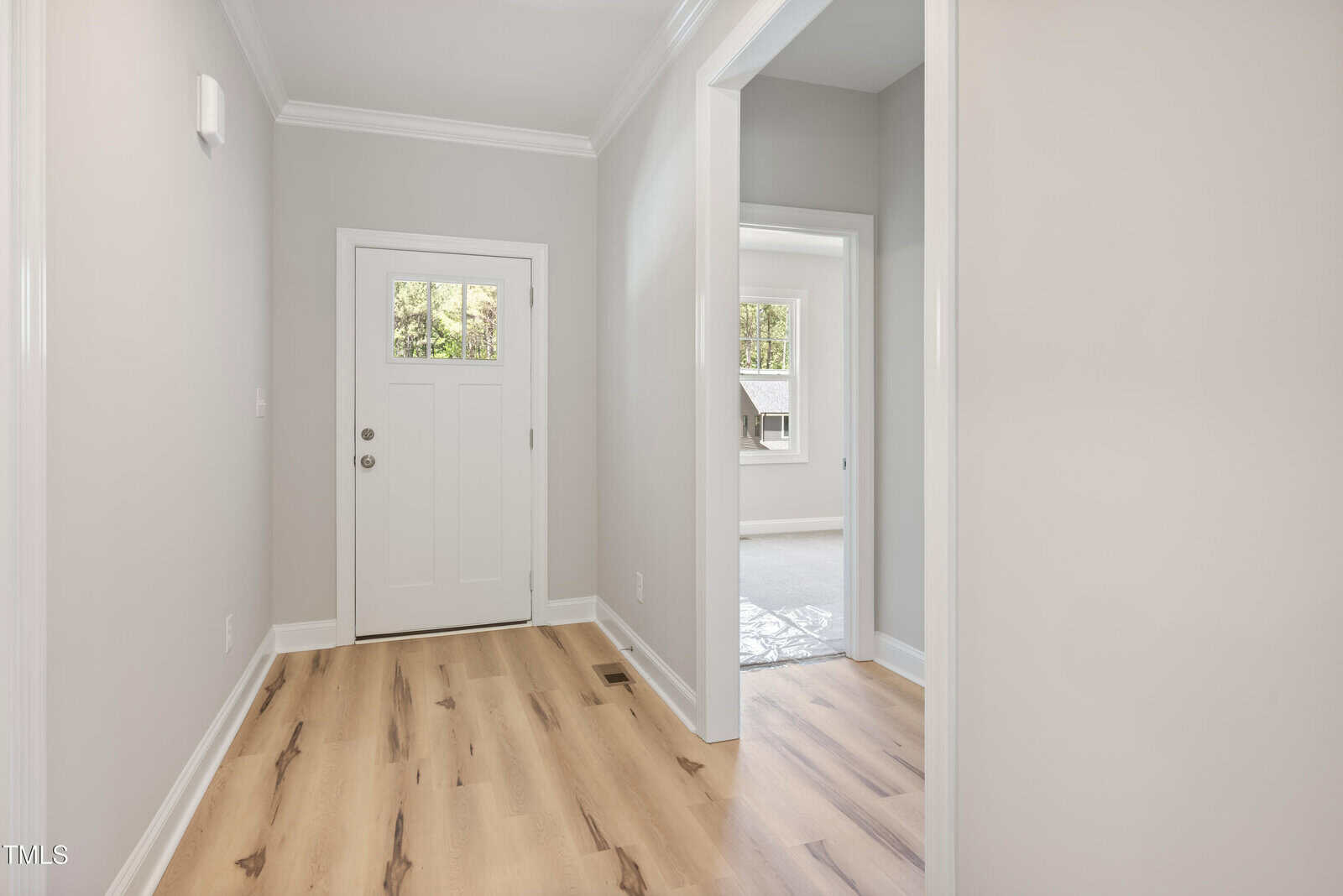 A bright, modern entryway with light wood floors, beige walls, white trim, and a white front door with a window. A hallway leads to another room with large windows and natural light.