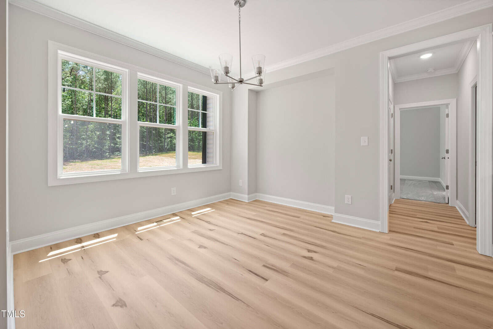 Bright, empty room with light wood flooring, neutral walls, three large windows, and a modern chandelier; open doorway leads to a hallway.