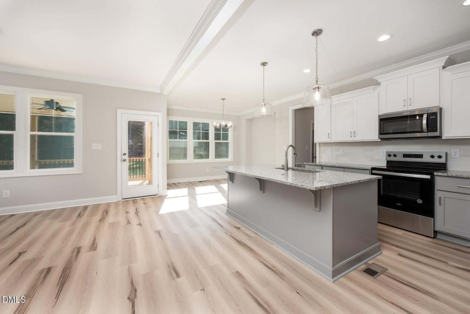 Bright, modern kitchen and dining area with light wood flooring, white cabinets, stainless steel appliances, a gray island with a sink, pendant lighting, and large windows letting in natural light.