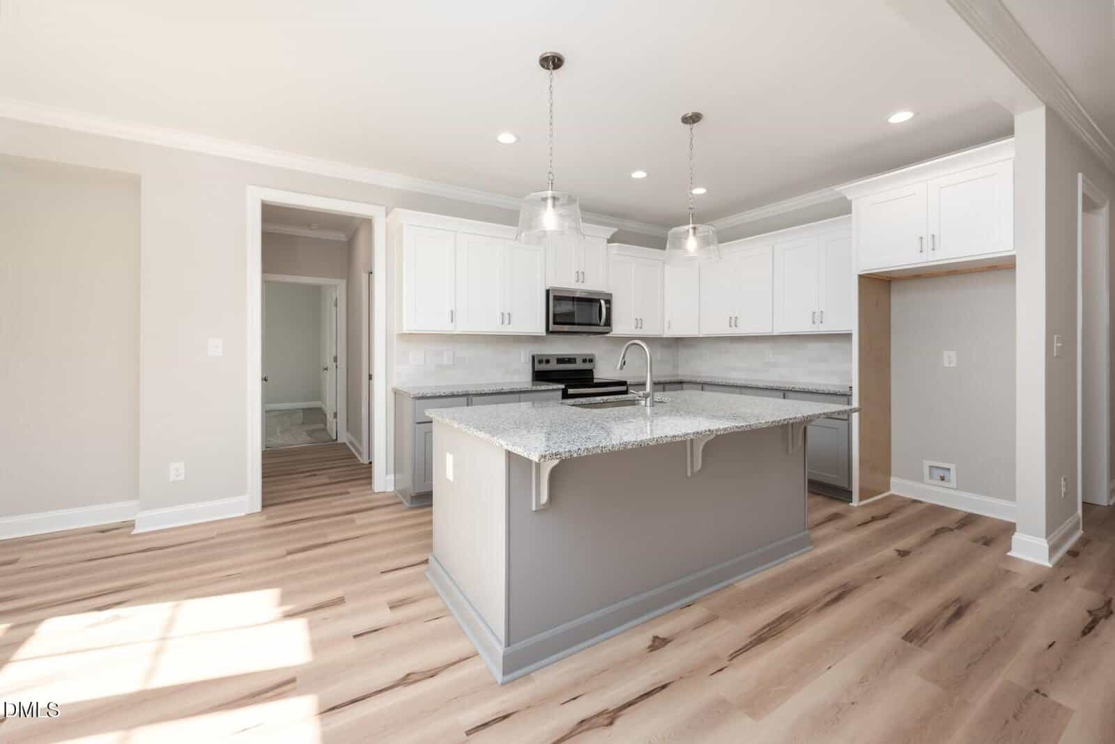 Modern kitchen with white cabinets, stainless steel appliances, a gray island with granite countertop, pendant lights, light wood flooring, and neutral walls. Sunlight streams in from a window on the left.