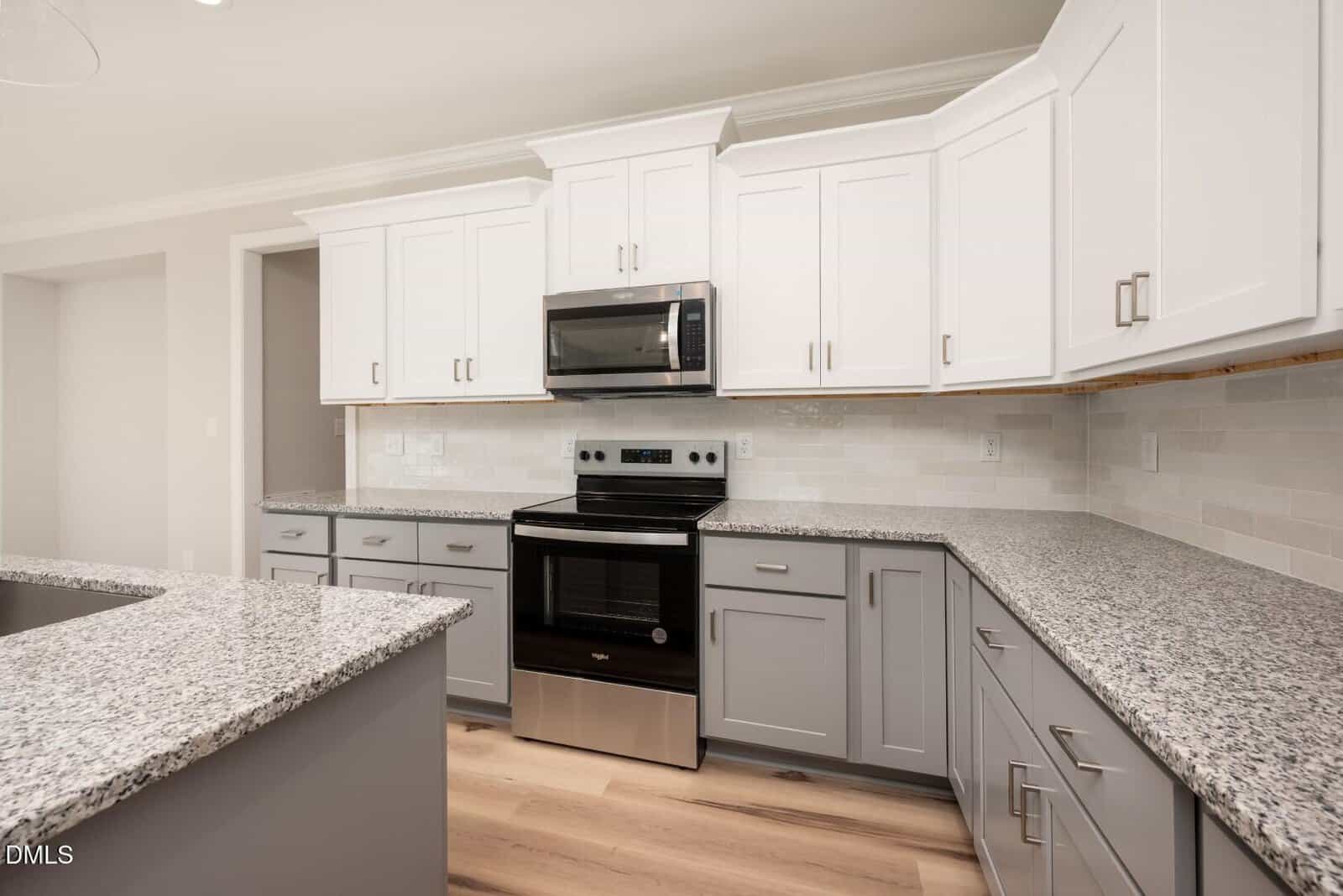 Modern kitchen with white upper cabinets, gray lower cabinets, granite countertops, stainless steel oven and microwave, light wood floors, and a neutral tile backsplash. The space is clean, bright, and spacious.