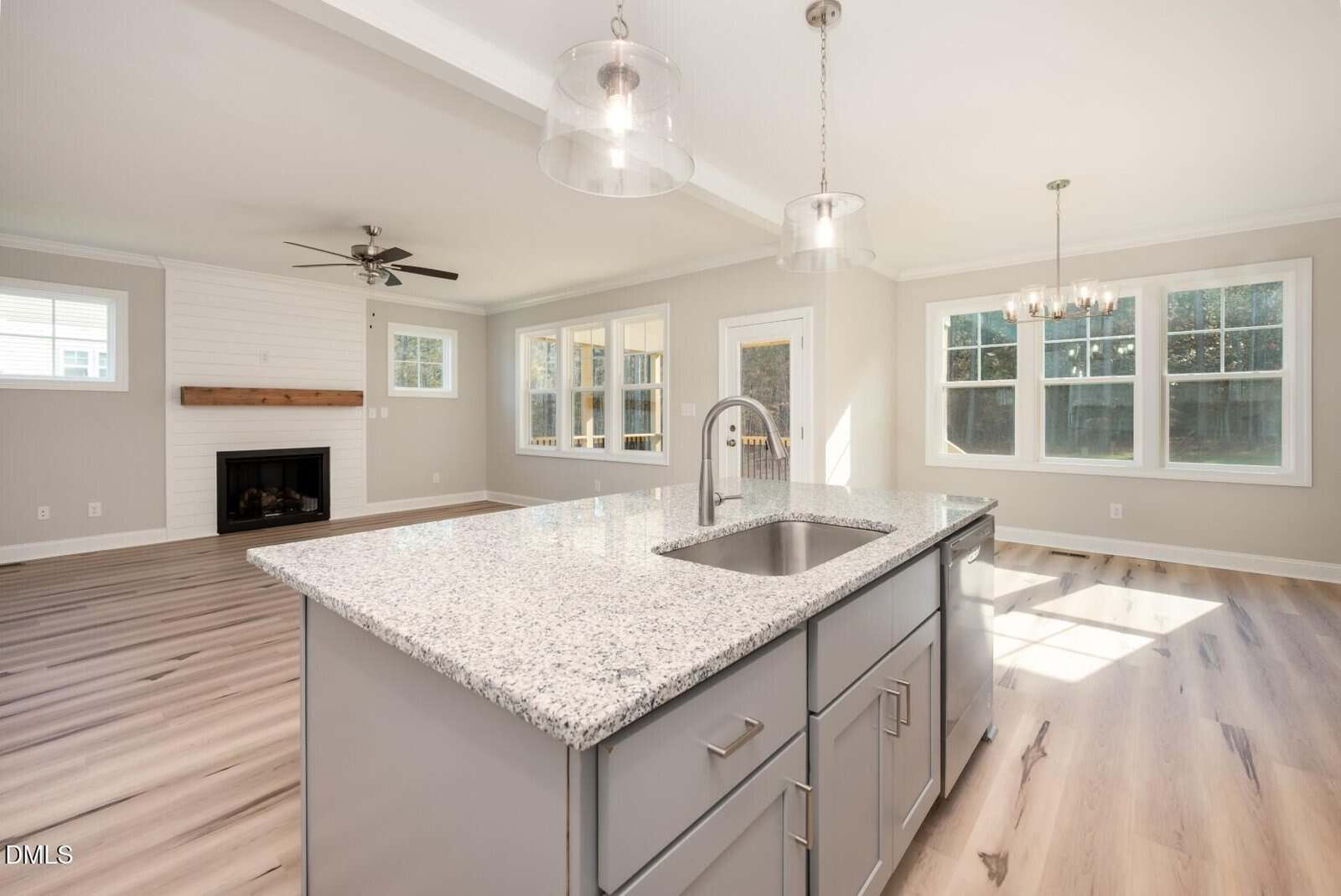 Bright, modern kitchen with a granite island and sink, open to a living area with a fireplace, ceiling fan, and large windows letting in natural light. Light wood floors throughout.