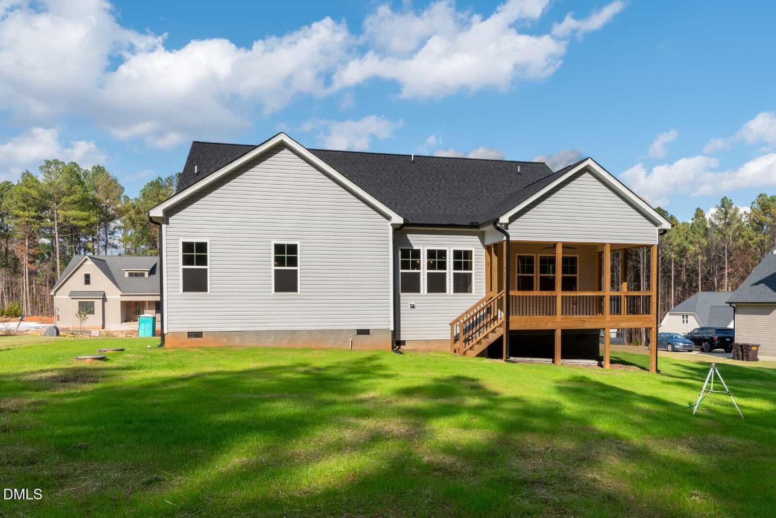 A light gray house with a black roof, white trim, and a screened-in porch, sits on a grassy lawn with trees in the background under a partly cloudy sky. Other houses are visible nearby.