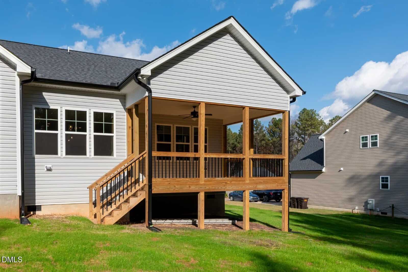 A modern house with light gray siding features a covered wooden porch with railings and stairs leading down to a green lawn. Another similar house is visible nearby under a clear blue sky.