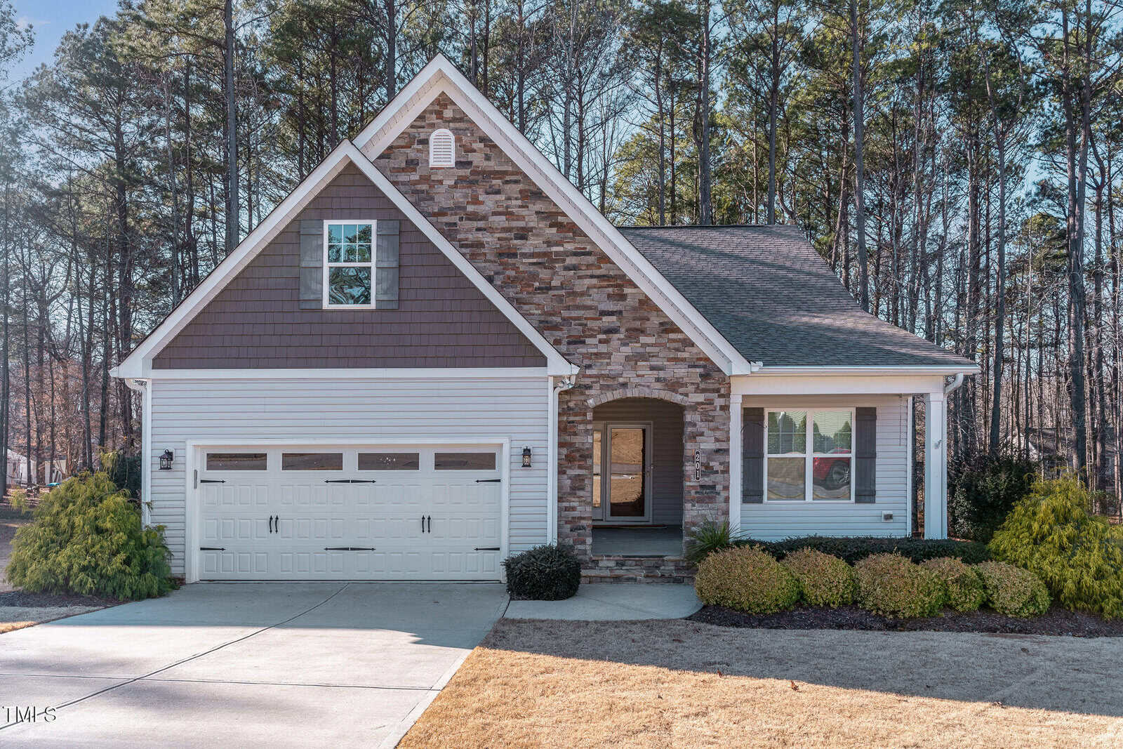 A single-story suburban house with a stone and shingle front, attached two-car garage, gabled roof, and a small covered porch. The yard has some shrubs, and there are tall trees in the background.