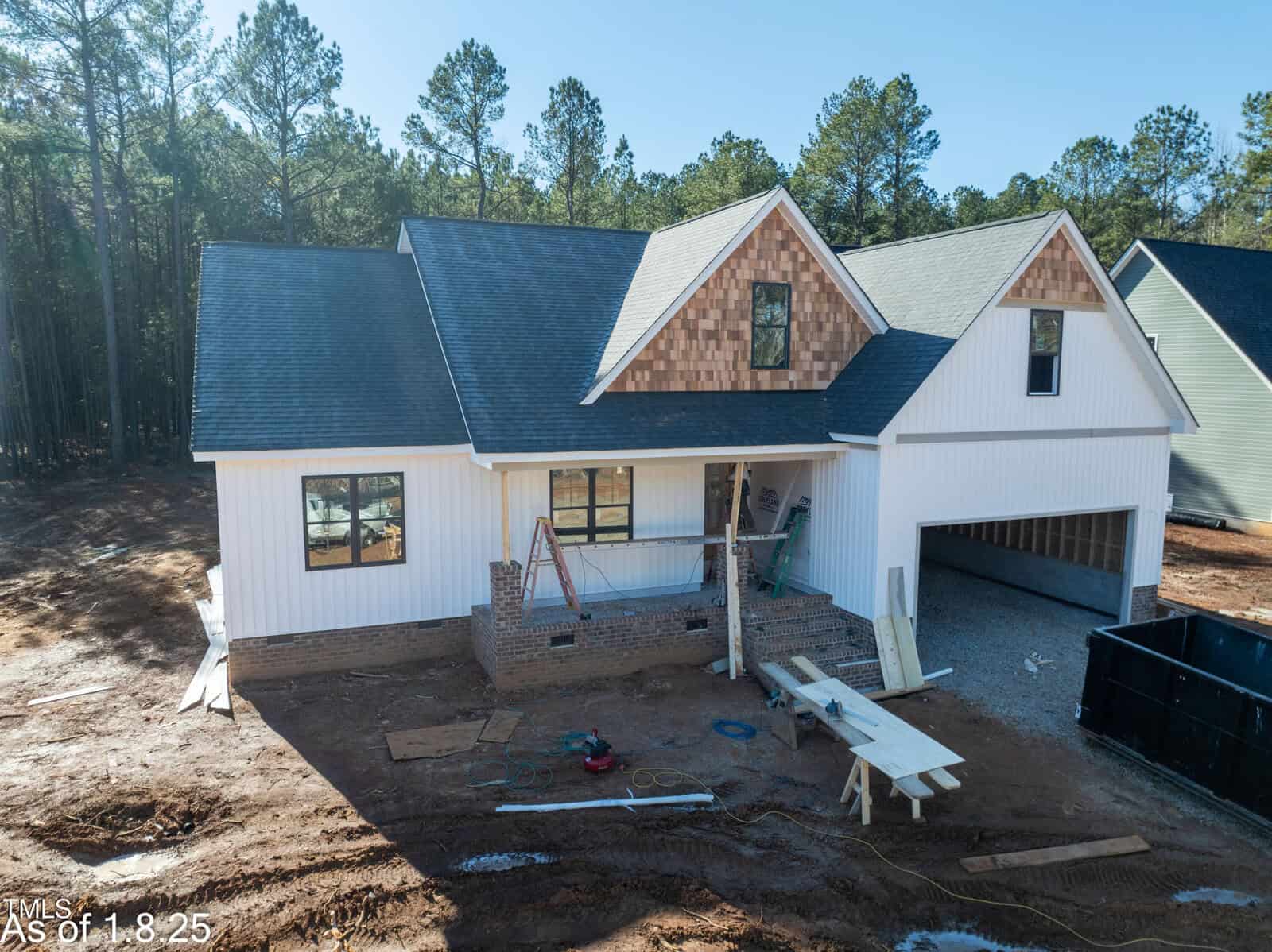 A white house under construction with exposed brickwork on the front porch, wood planks, and construction materials in the yard; surrounded by pine trees, with a two-car garage on the right. Timestamp reads "AS of 1.8.25" in the corner.